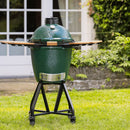 Green ceramic barbecue grill on a stand outdoors with a garden background